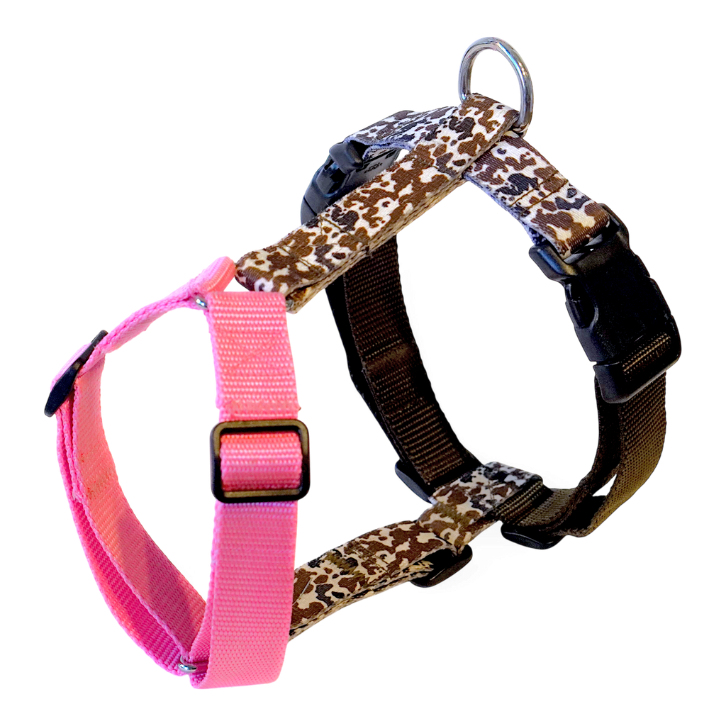 A Y-shape anatomical dog harness featuring a brown and beige cow print pattern with black plastic buckles, a silver metal ring, and a contrasting bright pink adjustable strap.