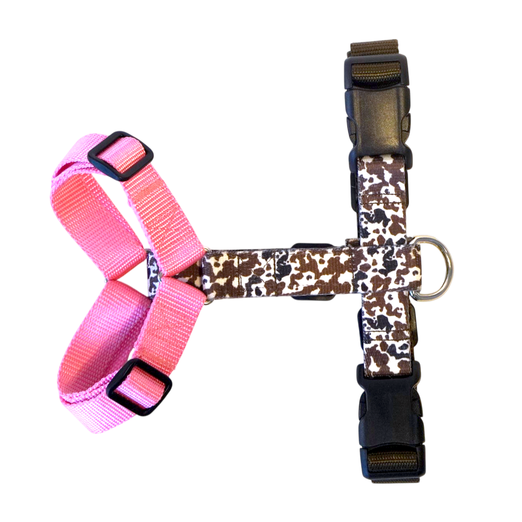 A Y-shape anatomical  dog harness with a pink left side and cow patterned right side, featuring black plastic buckles and a silver metal ring.